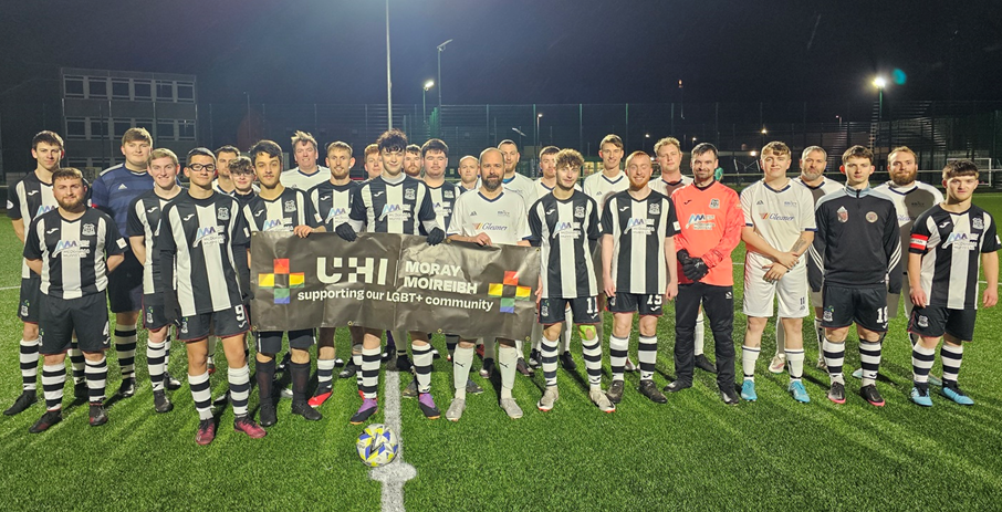 A team photo of all participants of the UHI Football vs Homophobia match. They are wearing their football kits and are holding a sign that reads 