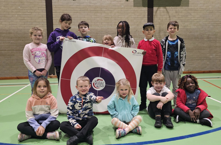 A group photo of Primary School pupils taking part in the Curling programme. They are in the UHI Moray Games Hall, some are sat on the floor whilst others are stood, holding a pieces of Curling equipment.