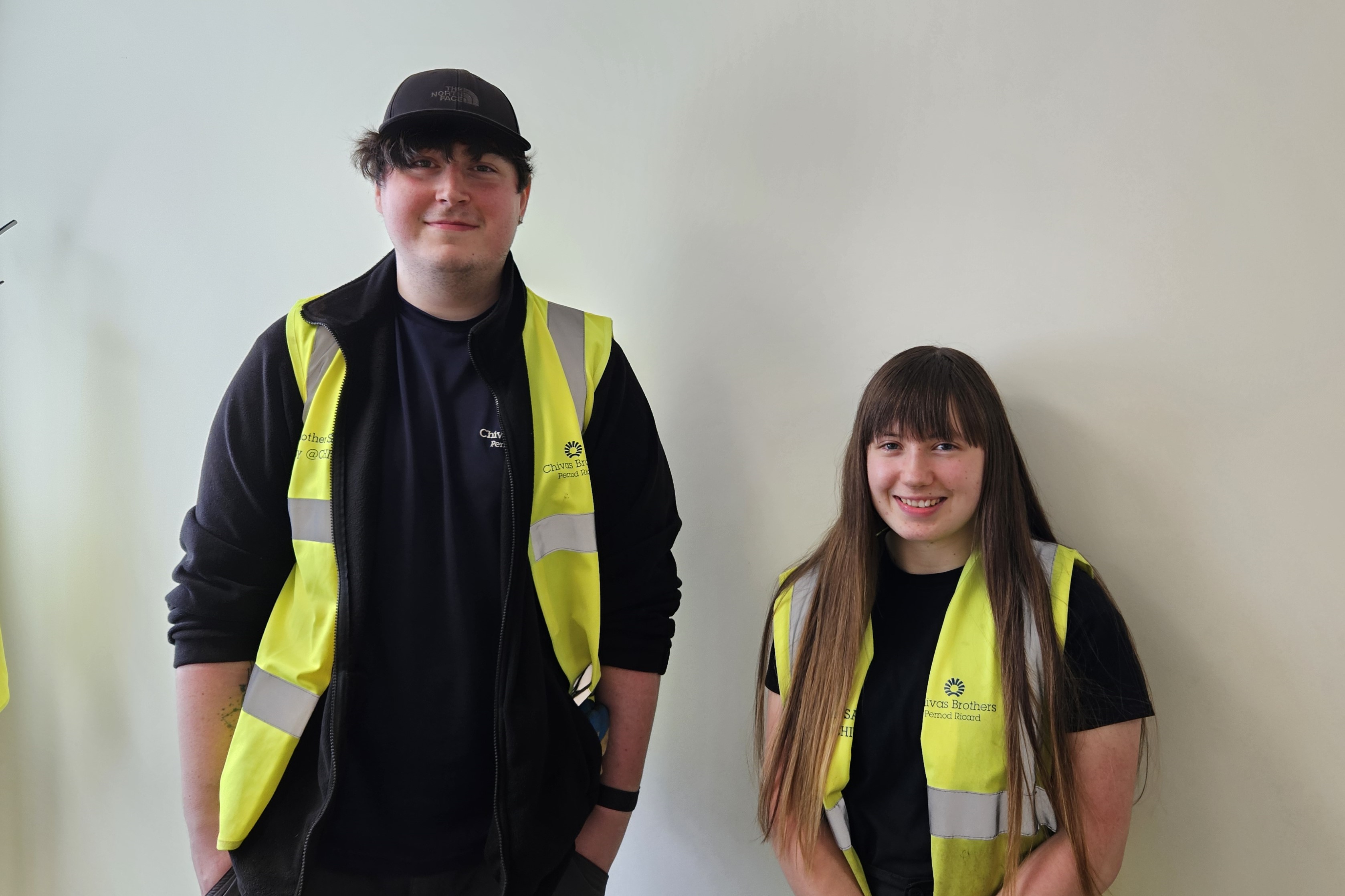 Photograph of employability clients, Dylan and Lucy, smiling at the camera and wearing hi-viz jackets in their place of work - Chivas