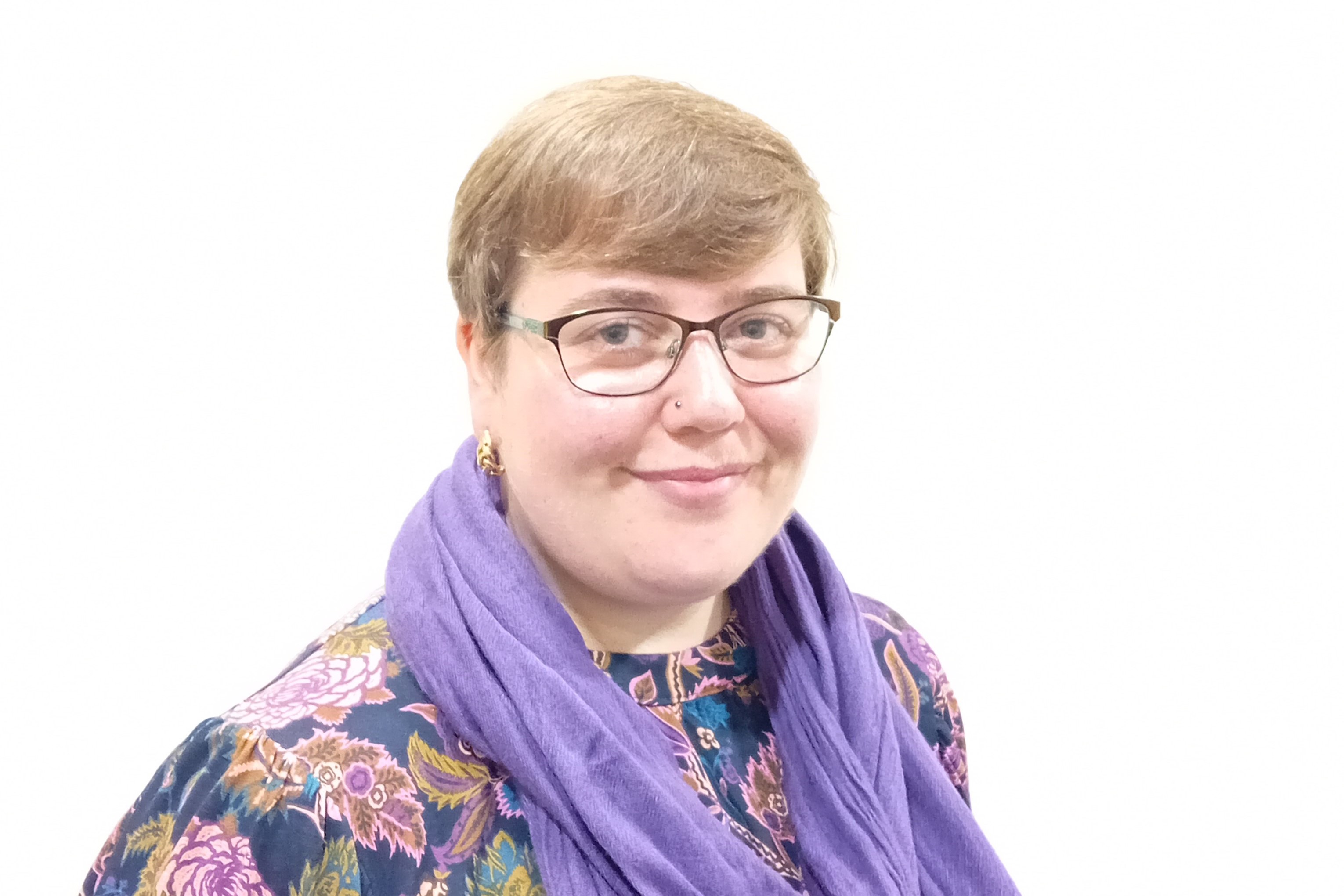 Photograph of employability client, Rebecca, smiling at the camera. She has short pixie cut hair, glasses, is wearing a purple scarf and a multicolour floral patterned top.