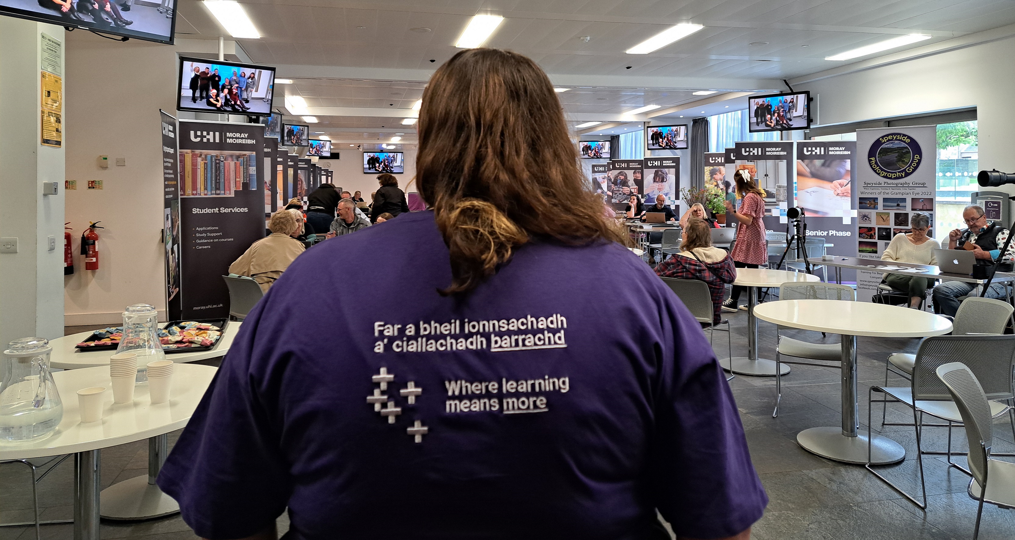 Photo of a person with their back faced to us looking into our AGBC Conference Room. The person is wearing a purple t-shirt which says Where Learning Means More in both English and Gaelic